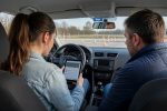 Žákyně autoškoly s tabletem a zkušebním testem sedí za volantem vedle instruktora na cvičné ploše